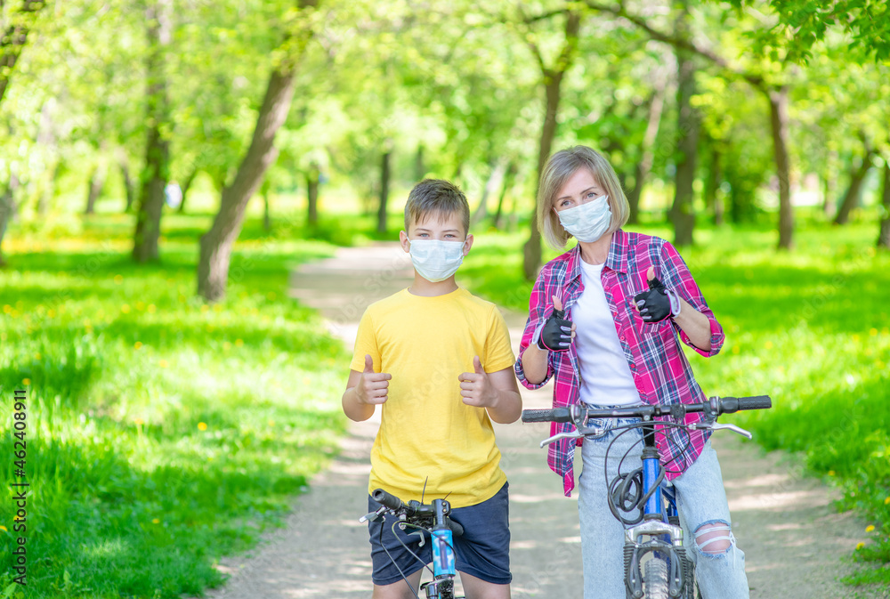 Mother and her young son wearing medical protective masks ride bikes in a summer park and show thumbs up gesture. Empty space for text Mother and her young son wearing medical protective masks ride bikes in a summer park and show thumbs up gesture. Empty space for text