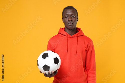 Young african american man guy football player in red streetwear hoodie posing isolated on yellow wall background. People emotions, sport leisure lifestyle concept. Playing football hold soccer ball. © ViDi Studio