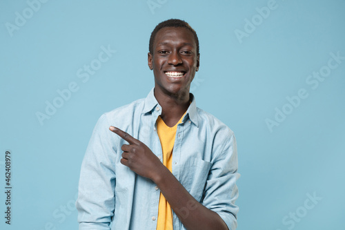 Smiling young african american man guy in casual shirt, yellow t-shirt posing isolated on blue background studio portrait. People lifestyle concept. Mock up copy space. Pointing index finger aside.