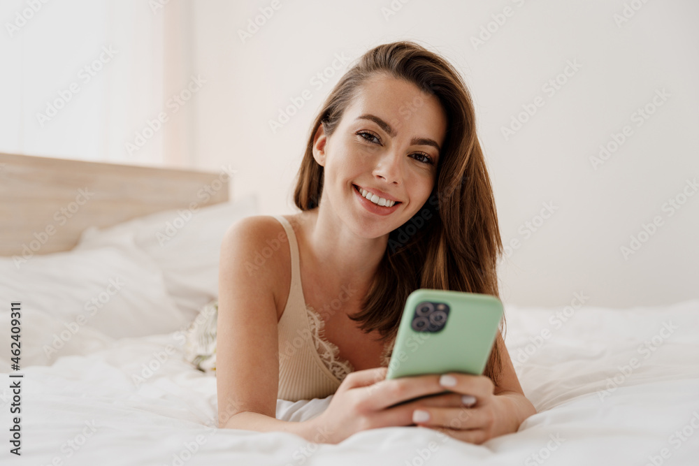 Fun young woman in casual casual clothes lying on her stomach hold in hand use mobile cell phone look camera rest relax spend time in bedroom lounge home in own room house be lost in reverie good day. Fun young woman in casual casual clothes lying on her stomach hold in hand use mobile cell phone look camera rest relax spend time in bedroom lounge home in own room house be lost in reverie good day.