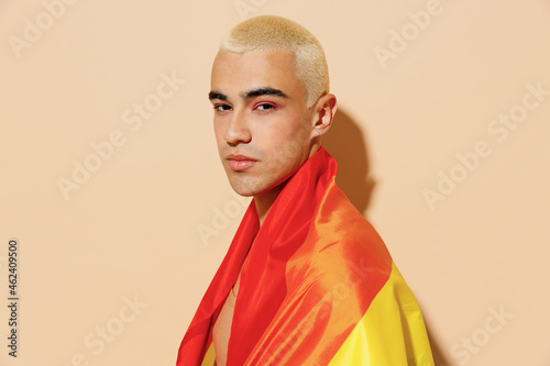 Side view sexy serious young blond latin gay man 20s with make up in beige tank shirt wrapped in rainbow flag look camera isolated on plain light ocher background studio People lgbt lifestyle concept.