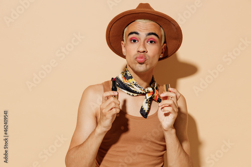 Young trendy sexy blond latin gay man 20s with make up duckface in beige tank shirt neck scarf hat hold lipstick isolated on plain light ocher background studio portrait People lgbt lifestyle concept. photo