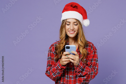 Merry smiling pensive fun young Santa woman 30s wears shirt Christmas hat hold use mobile cell phone typing browsing chatting send sms isolated on plain pastel light violet background studio portrait.