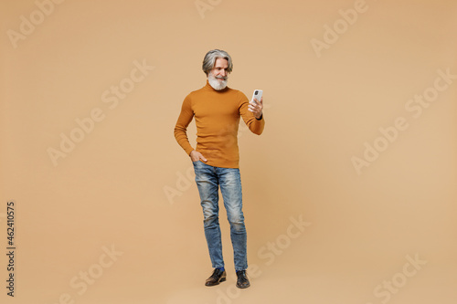 Full body elderly gray-haired mustache bearded man 50s in mustard yellow turtleneck shirt hold in hand use mobile cell phone isolated on plain pastel beige background studio. People lifestyle concept.