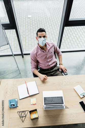 Top view of abian businessman in medical mask looking at camera near devices and notebook in office photo