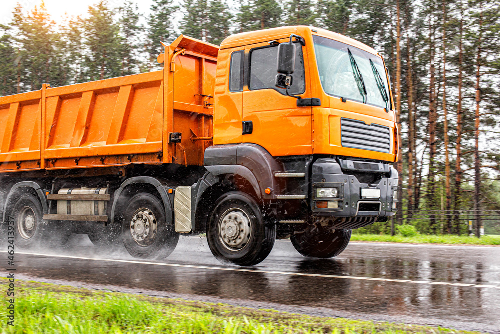 An orange 4-axle cargo dump truck with a load of 40 tons drives on a wet road in summer, in the background. Bulk cargo transportation, bad weather