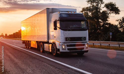 White truck with refrigerated semitrailer transport frozen food on the highway in the evening against the backdrop of sunset. Transportation of perishable goods