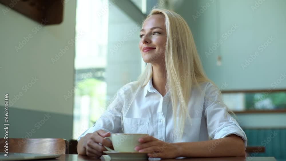 Close-up of smiling attractive young blonde woman drinking with cup of hot coffee and looking outside through window in coffee shop. Gorgeous happy lady sitting in cafe with cup of tea or coffee. Close-up of smiling attractive young blonde woman drinking with cup of hot coffee and looking outside through window in coffee shop. Gorgeous happy lady sitting in cafe with cup of tea or coffee.