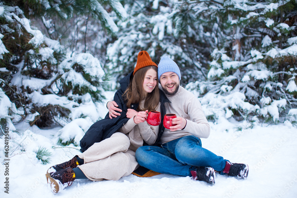 Fototapeta premium A loving girl and a guy sit in the snow and drink tea from red cups in winter against the background of a snow-covered coniferous forest