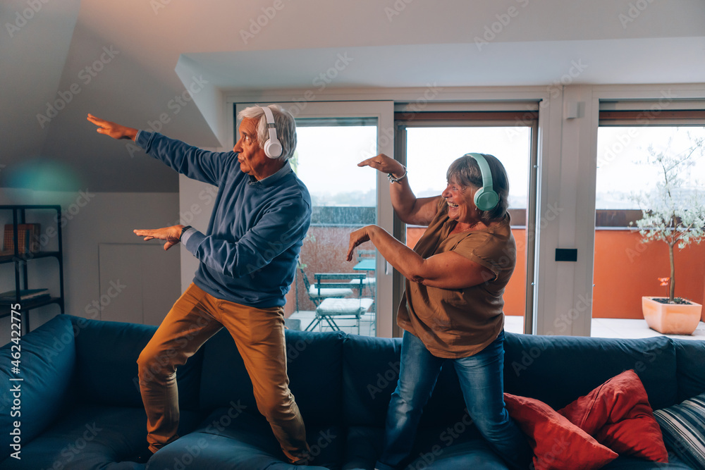 happy active senior couple plays and dances on sofa at home with headphones listening to music - obrazy, fototapety, plakaty 