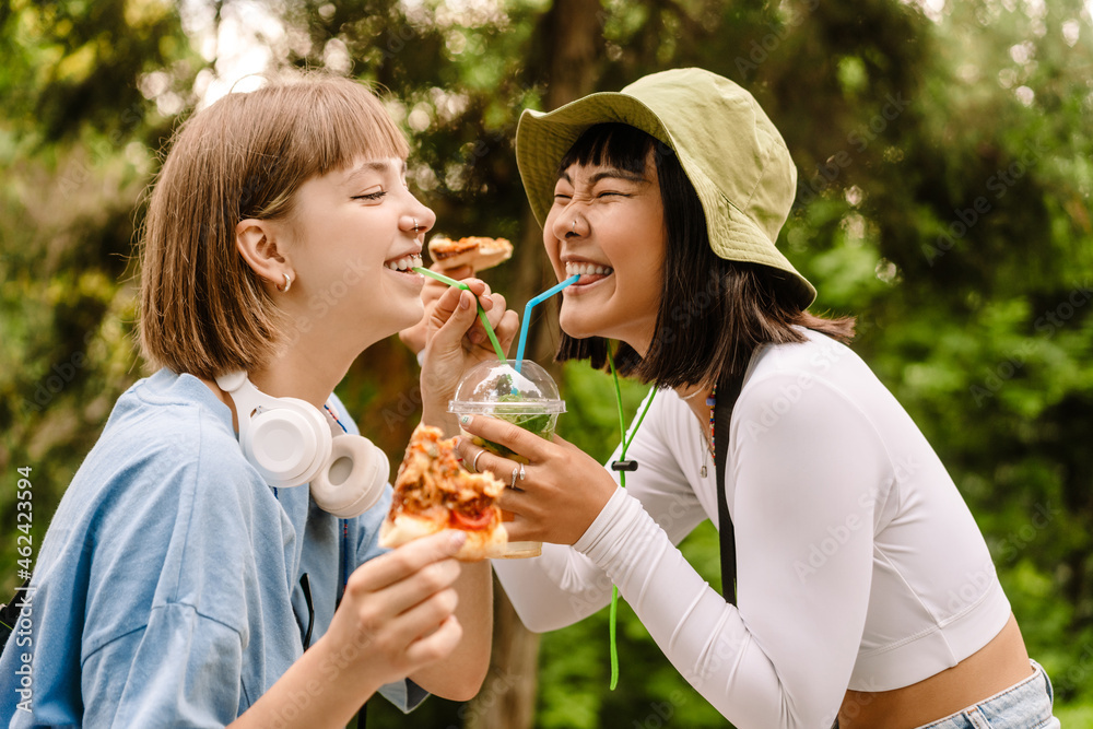 Fototapeta premium Multiracial two women drinking soda and eating pizza while resting in park