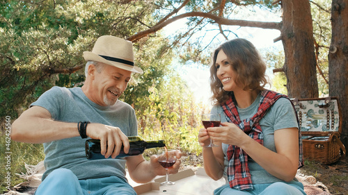A married couple is relaxing in nature and drinking wine, and having fun talking while sitting in the woods on a picnic on a summer day.