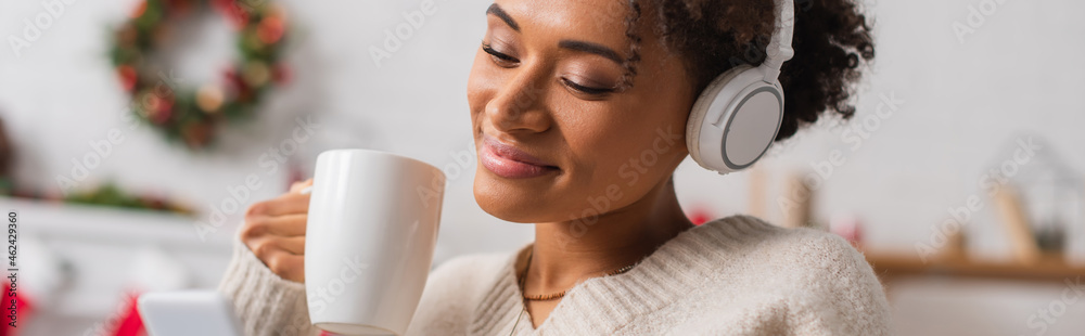 Happy african american woman in headphones holding cup while looking at smartphone during christmas at home, banner - obrazy, fototapety, plakaty 