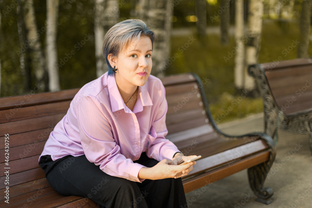 Teenage girl with her pursed lips is sitting on a bench in park. Teenager, 18 year old, is holding a smartphone in his hands and looking at camera. Teenage girl with her pursed lips is sitting on a bench in park. Teenager, 18 year old, is holding a smartphone in his hands and looking at camera.