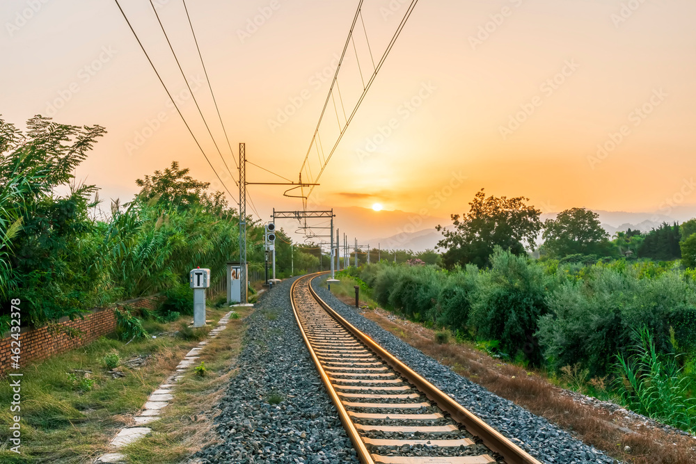 old evening railroad leading to a sunset glow in mountains with green bushes and cjlorful cloudy sku on the background