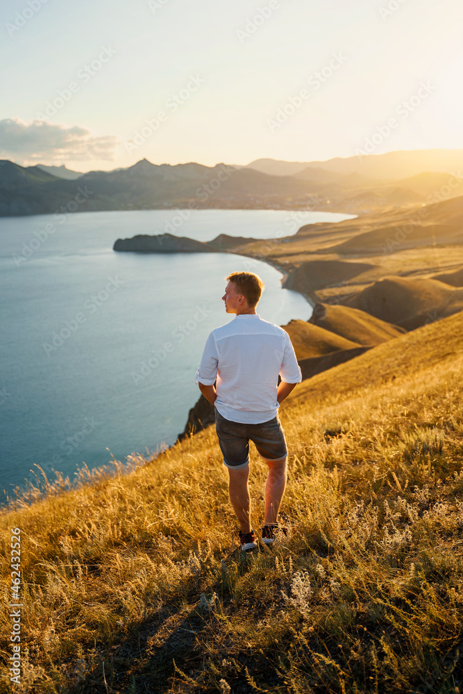 A young man travels along the mountain coast at sunset. Rear view A man stands on a mountainside and enjoys the view. The concept of freedom and achievement of goals.