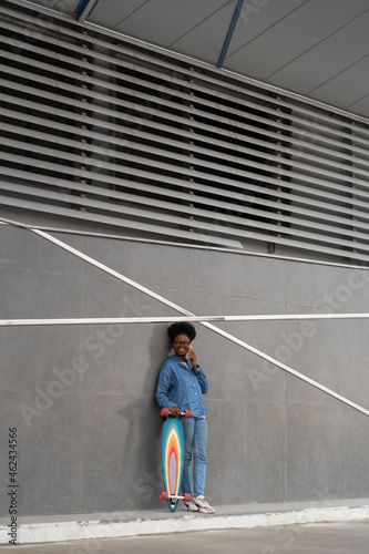 African american hipster girl student skater make phone call standing near wall of building with longboard outdoors. Young woman skateboarder talking on cellphone, resting. 