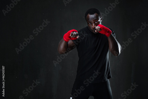 Sporty man during boxing hit. African american muscular athlete in sport uniform and red bandage over black background. Strength, fighting and motion project.