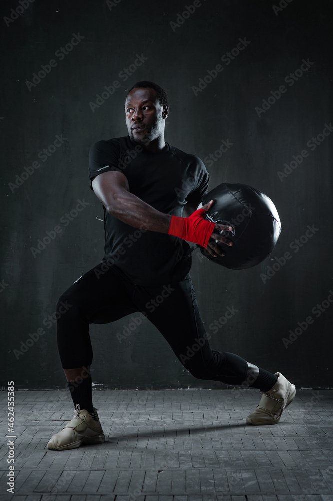 Functional strength exercises for boxers. African American muscular fitness man is working out with medicine ball over black background. Boxing strength and conditioning workout project. Copy space.
