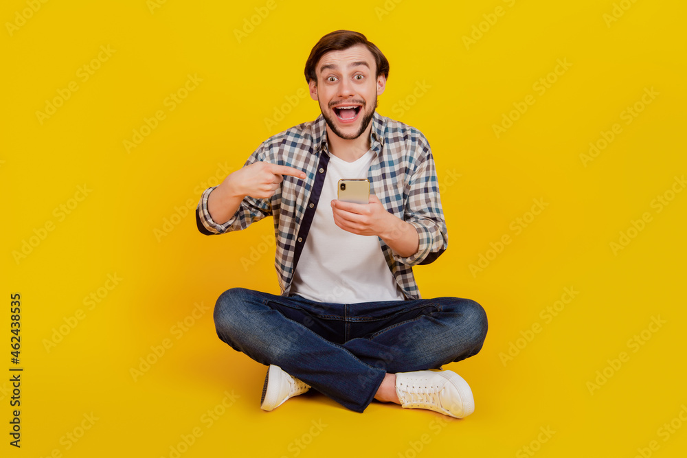 Full length photo of excited cheerful man sit floor point finger cellphone advert recommend promo isolated yellow color background Full length photo of excited cheerful man sit floor point finger cellphone advert recommend promo isolated yellow color background