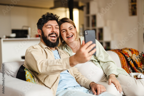 White couple laughing and using cellphone while sitting on couch at home