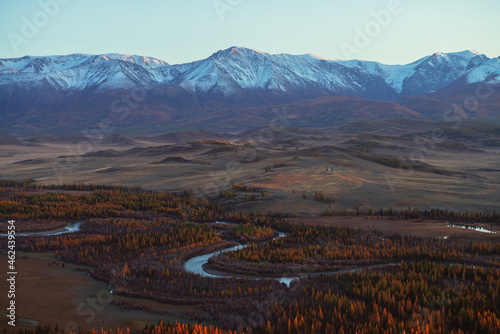Awesome autumn landscape with mountain river in forest valley and great snow-covered mountain range in red sunset sunshine. Spectacular view from hill to snowy mountains and autumn valley in sunlight. © Daniil
