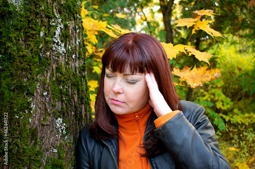 Photography Red-haired middle-aged woman is having a headache or migraine and is leaning on a tree in the park