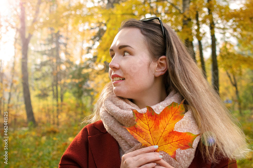Young woman wearing warm scarf and coat is holding orange maple leaf and smiling in the park. Enjoying sunny weather