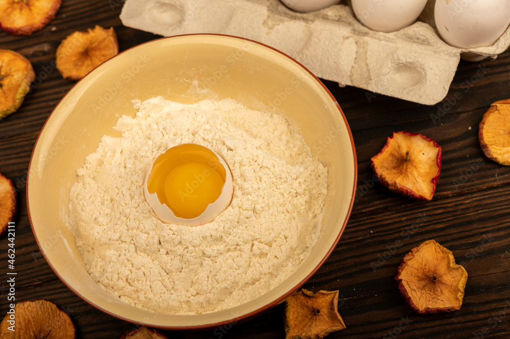 A broken white chicken egg in a bowl with wheat flour, dried apple chips and several whole eggs in a tray made of white cardboard, close-up selective focus.