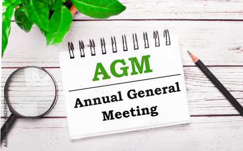 On a light wooden background, a magnifying glass, a pencil, a green plant and a white notebook with text AGM Annual General Meeting. Business concept photo