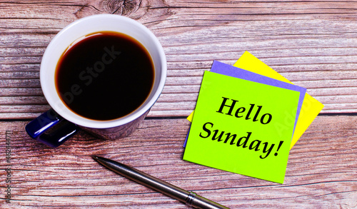 On a wooden table - a blue cup with coffee, a pen and colorful bright stickers with the text HELLO SUNDAY. View from above
