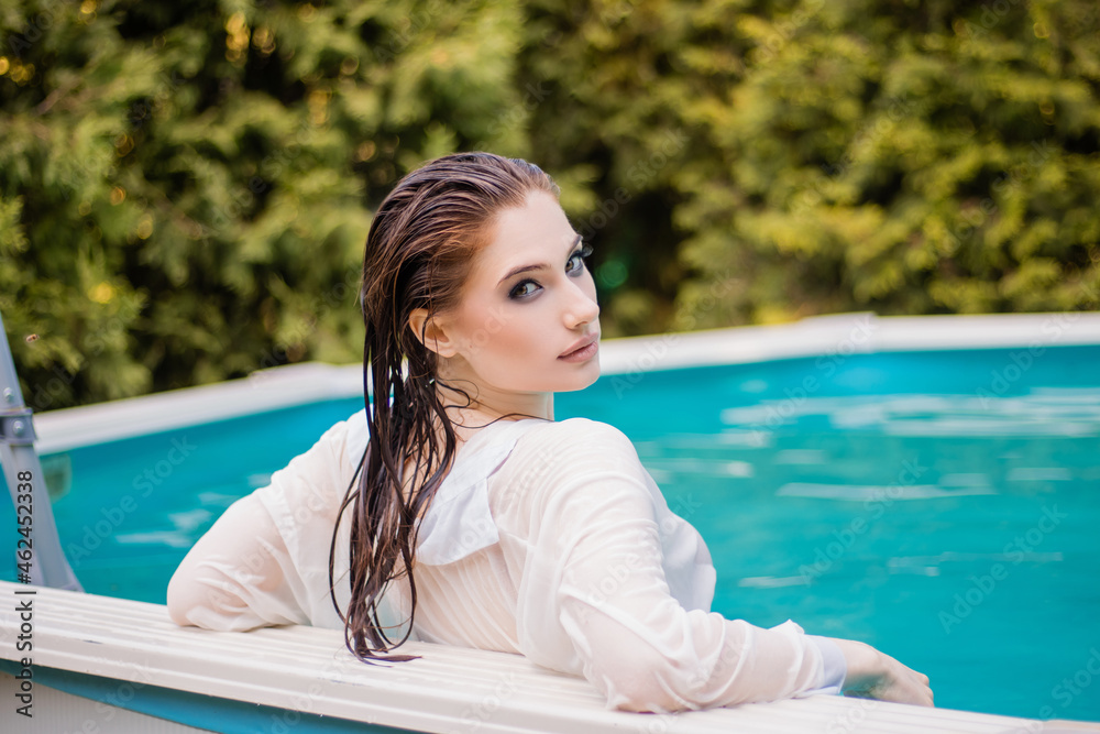Portrait of a beautiful girl with wet hair dressed in a white shirt standing chest-deep in water near the edge of the pool.
