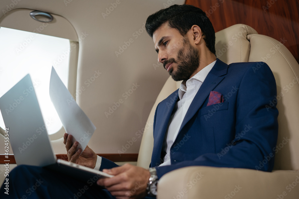 Handsome pensive middle eastern businessman using laptop, reading financial report sitting business class on plane. Rich confident entrepreneur flying luxury private jet, successful business concept Handsome pensive middle eastern businessman using laptop, reading financial report sitting business class on plane. Rich confident entrepreneur flying luxury private jet, successful business concept