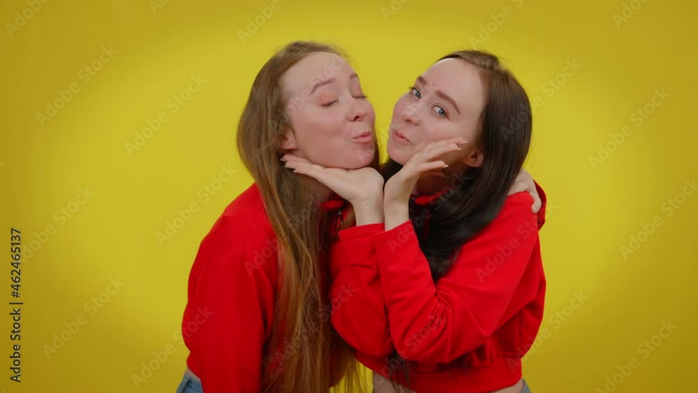 Funny millennial twin sisters grimacing kissing posing at yellow background. Portrait of carefree Caucasian young beautiful women having fun making faces looking at camera smiling Funny millennial twin sisters grimacing kissing posing at yellow background. Portrait of carefree Caucasian young beautiful women having fun making faces looking at camera smiling