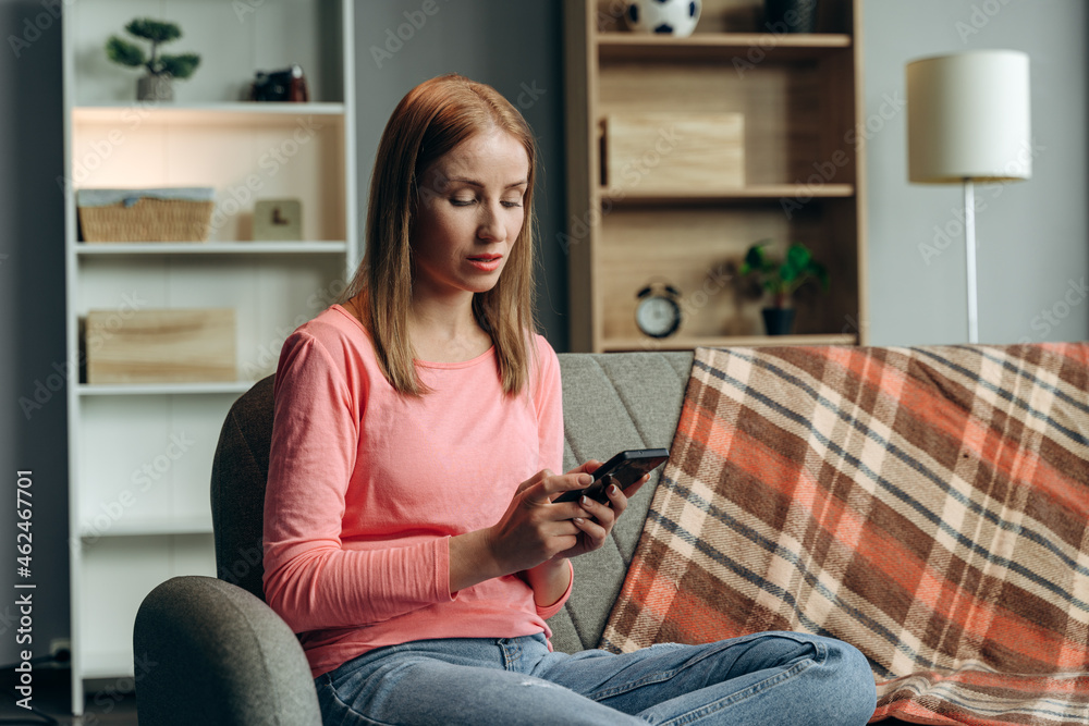 Ginger young concentrated woman at home using modern smartphone while sitting at the sofa. Online chat concept Ginger young concentrated woman at home using modern smartphone while sitting at the sofa. Online chat concept