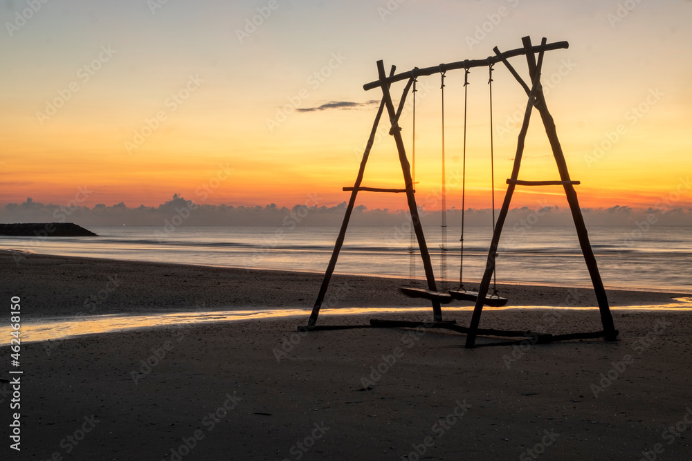 A large wooden swing on a sandy beach in the evening with a golden sky. A large wooden swing on a sandy beach in the evening with a golden sky.