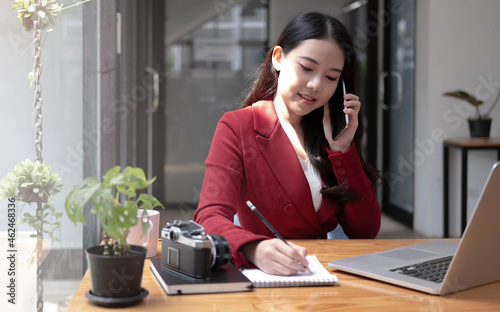 Young woman talking on mobile phone and writing notes while sitting at her desk. Asianfemale working in home office photo