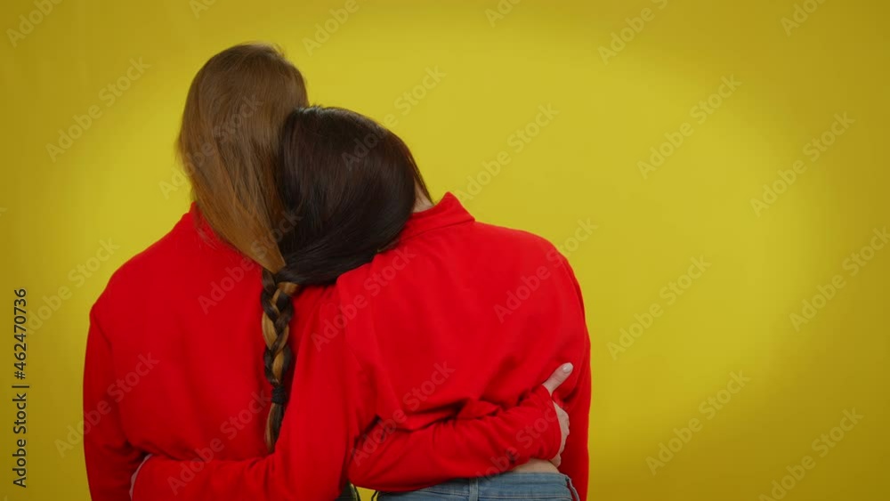 Back view twin sisters with red and black hair braided together standing at yellow background hugging. Concept of sisterhood unity and individuality Back view twin sisters with red and black hair braided together standing at yellow background hugging. Concept of sisterhood unity and individuality
