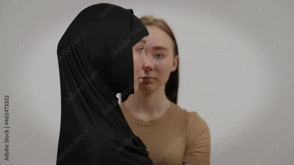 Side view woman in hijab posing at grey background with twin Caucasian sister at background. Confident religious millennial lady looking aside smiling. Confidence and religion concept Side view woman in hijab posing at grey background with twin Caucasian sister at background. Confident religious millennial lady looking aside smiling. Confidence and religion concept