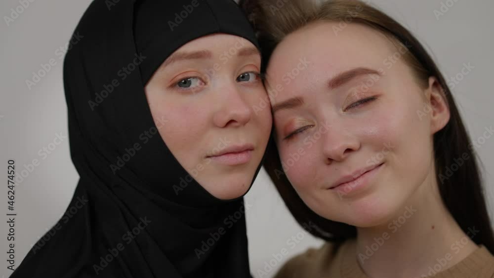 Close-up faces of twin sisters looking at camera smiling posing at grey background. Positive young Caucasian woman and sibling in black hijab face to face. Difference and individuality