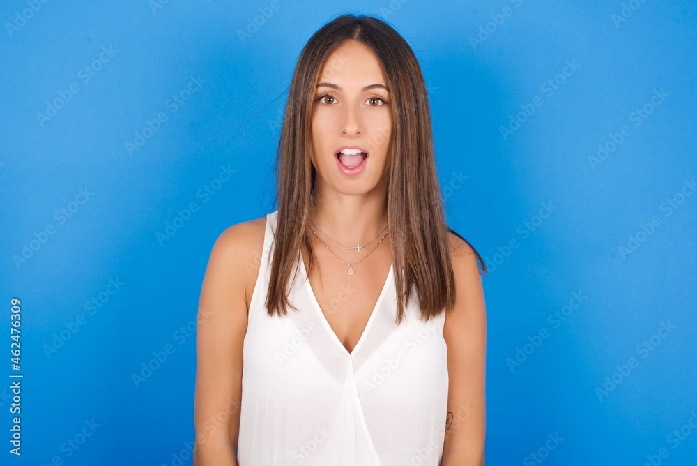 Young european brunette woman wearing white T-shirt on blue background having stunned and shocked look, with mouth open and jaw dropped exclaiming: Wow, I can't believe this. Surprise and shock