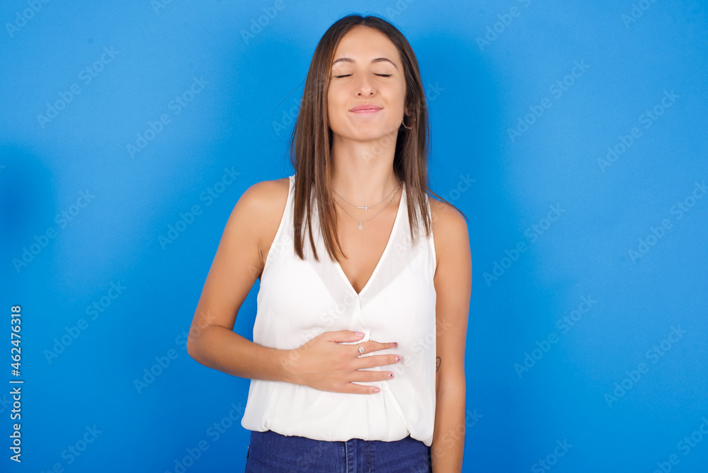 Satisfied smiling Young european brunette woman wearing white T-shirt , keeps hands on belly, being in good mood after eating delicious supper, demonstrates she is full. Pleasant feeling in stomach. Satisfied smiling Young european brunette woman wearing white T-shirt , keeps hands on belly, being in good mood after eating delicious supper, demonstrates she is full. Pleasant feeling in stomach.