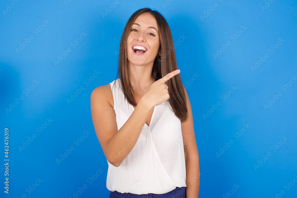 Young european brunette woman wearing white T-shirt on blue background pointing away and smiling to you. Look over there! Young european brunette woman wearing white T-shirt on blue background pointing away and smiling to you. Look over there!
