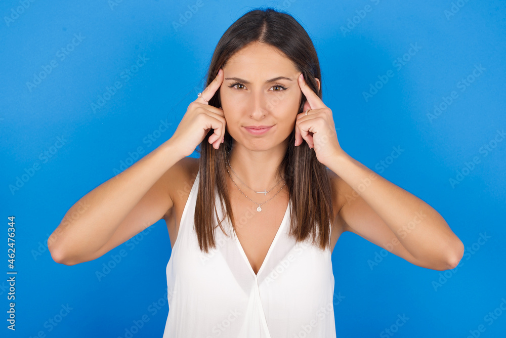 Young european brunette woman wearing white T-shirt on blue background concentrating hard on an idea with a serious look, thinking with both index fingers pointing to forehead. Young european brunette woman wearing white T-shirt on blue background concentrating hard on an idea with a serious look, thinking with both index fingers pointing to forehead.
