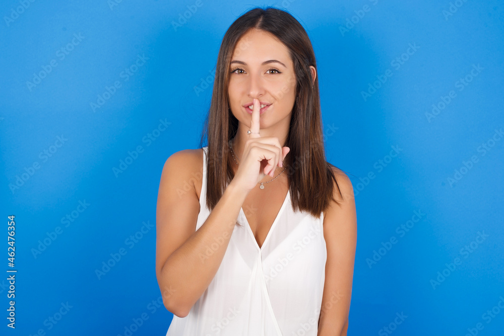 Young european brunette woman wearing white T-shirt on blue background makes hush gesture, asks be quiet. Don't tell my secret or not speak too loud, please! Young european brunette woman wearing white T-shirt on blue background makes hush gesture, asks be quiet. Don't tell my secret or not speak too loud, please!
