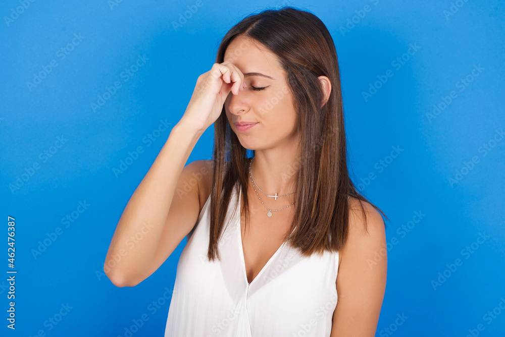 Very upset, Young european brunette woman wearing white T-shirt on blue background touching nose between closed eyes, wants to cry, having stressful relationship or having troubles with work