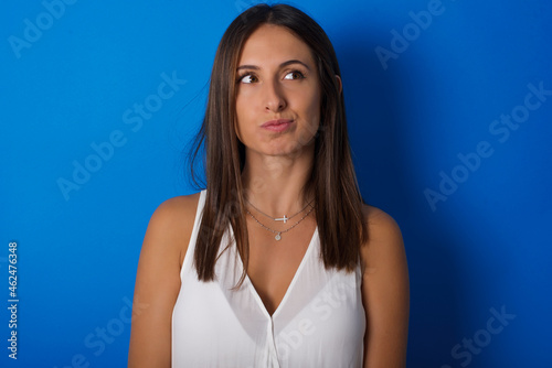 Young european brunette woman wearing white T-shirt on blue background, looks pensively aside, plans actions after university, imagines what to do Thinks over about new project.