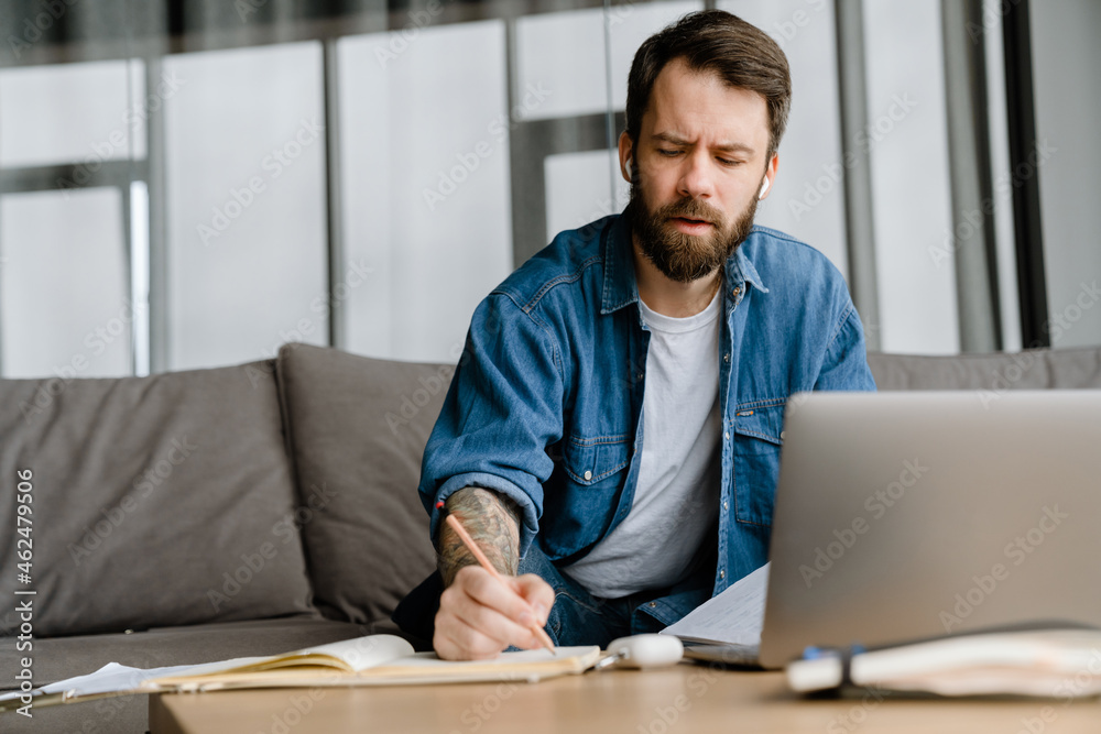 Bearded man writing down notes while making conference call on laptop Bearded man writing down notes while making conference call on laptop