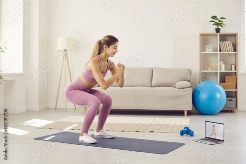 Fototapeta Naklejka Na Ścianę i Meble -  Woman using modern technology, watching video lesson and having fitness workout at home. Happy sporty lady standing on yoga mat and practicing squats during video call with online sports instructor