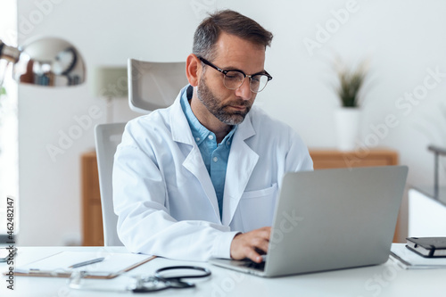 Mature concentrated attractive male doctor working with her computer in medical consultation.